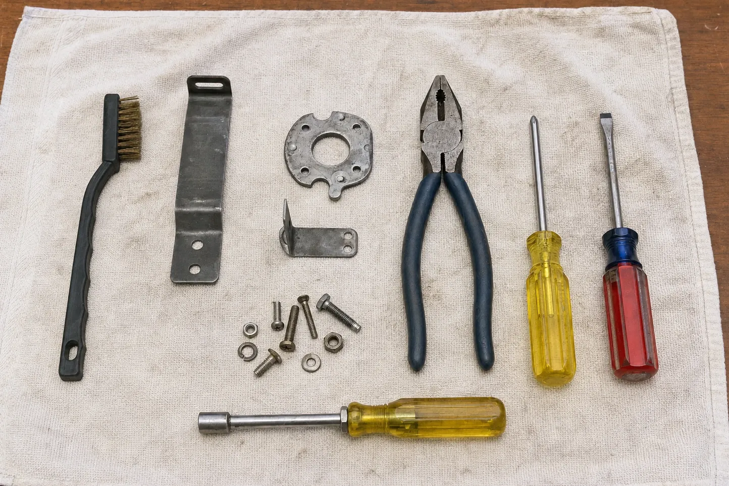Round burner mounting collar and an angle bracket between a wire brush, lineman pliers, a nut driver and loose bolts on a pale cloth
