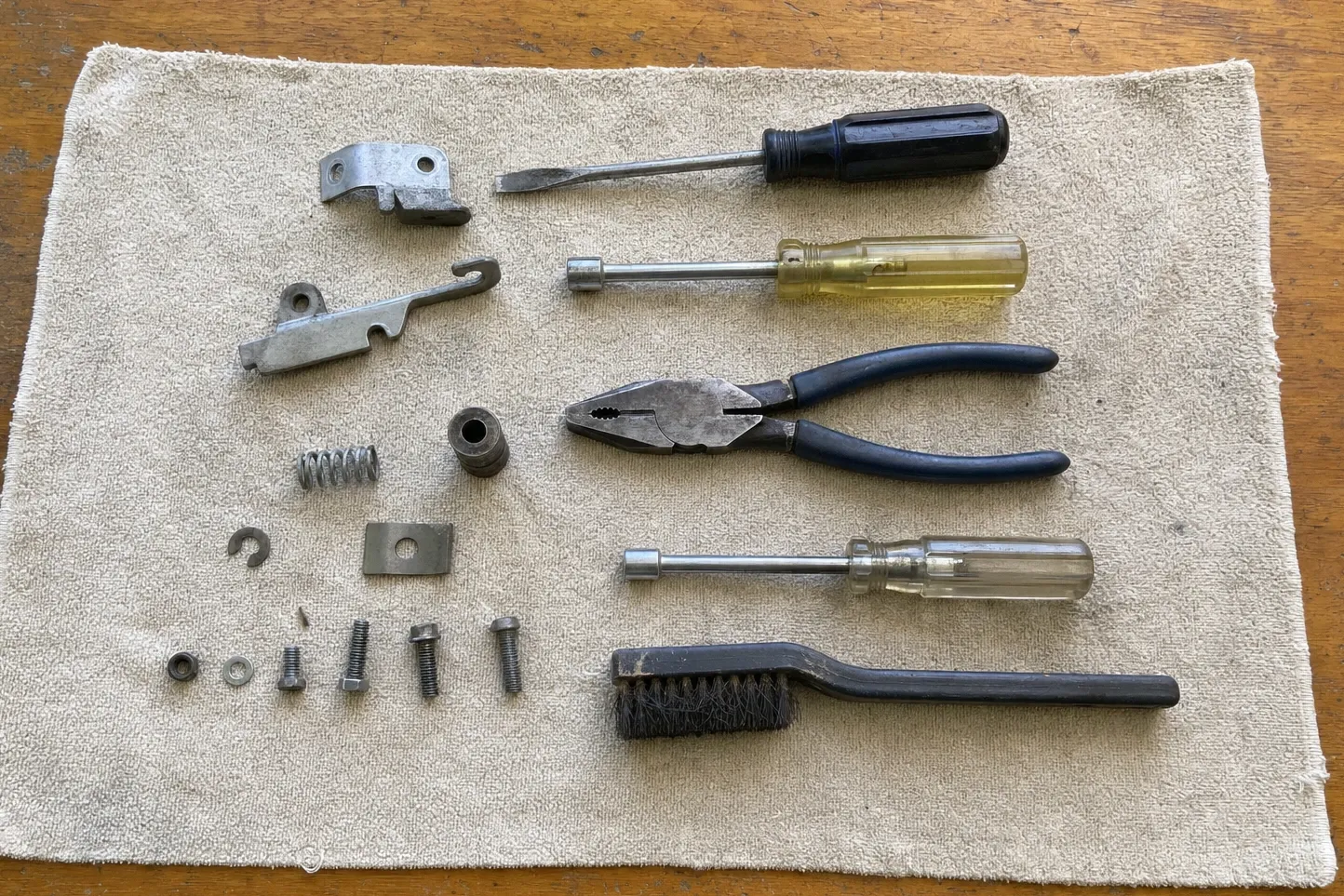 Latch bracket and a hooked latch lever with a coil spring, a bushing and a C-clip, laid beside drivers, pliers and a wire brush on burlap