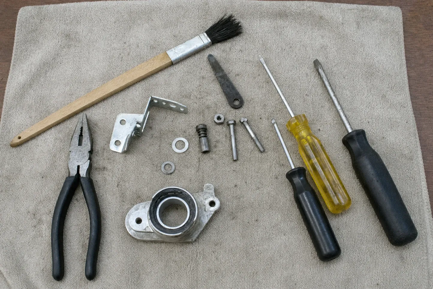 Bearing housing with its collar and seal beside a valve spindle, a bright hinge bracket, a long parts brush and three screwdrivers