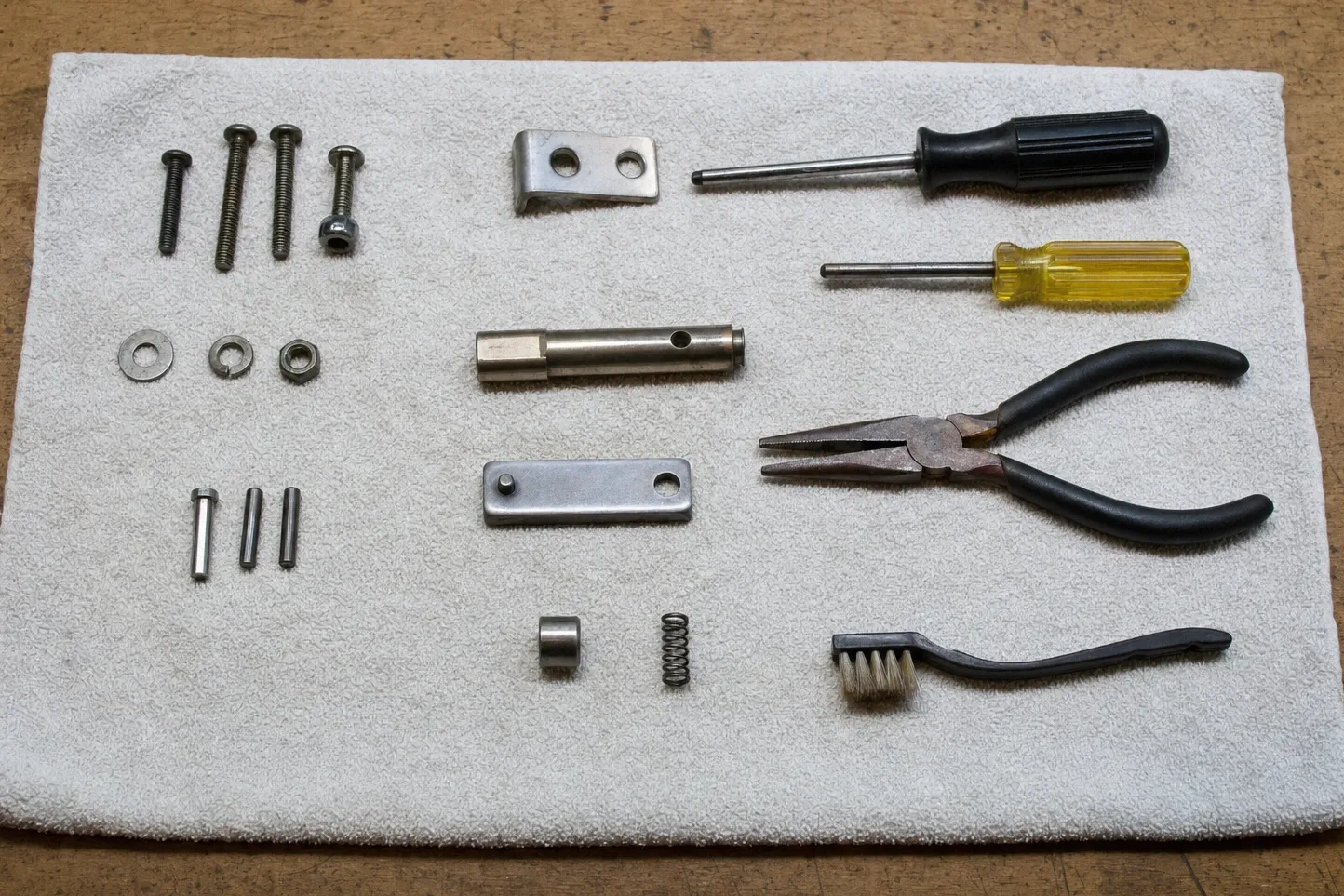 Machine screws, washers, three roll pins, two flat link plates, a drilled spindle and a small spring, with an awl driver and pliers alongside